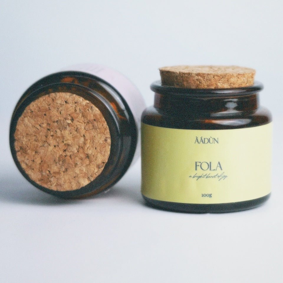 An amber glass jar with a cork lid and a pale yellow label reading 'AADUN FOLA', shown next to the open jar lid on a light gray background.