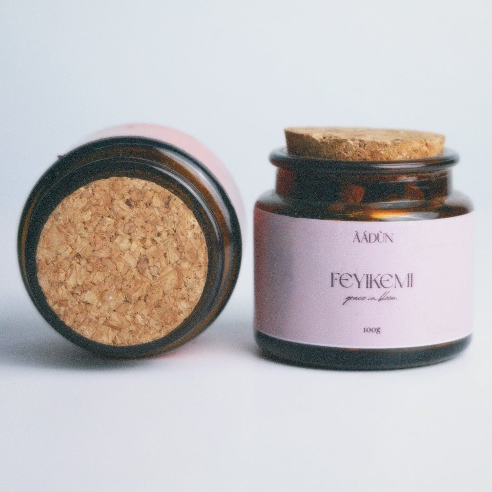 Two amber glass jars with cork lids on a light gray background. One jar is lying on its side showing the cork seal, and the other is standing upright with a pale pink label reading 'AADUN' and 'FEYIKEMI'.