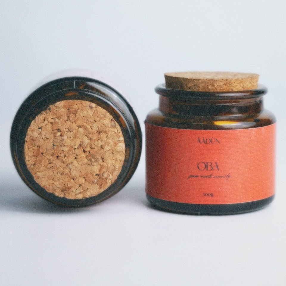 Two amber glass jars with cork lids on a light gray background. One jar is open, showing the textured cork insert, and the other is closed with a terracotta orange label that reads 'AADUN' and 'OBA'.