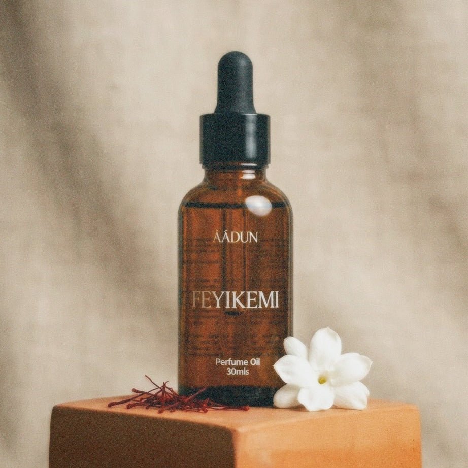 A 30ml amber glass dropper bottle of AADUN Feyikemi perfume oil, displayed on a terracotta block with a white flower and saffron threads against a textured beige background.