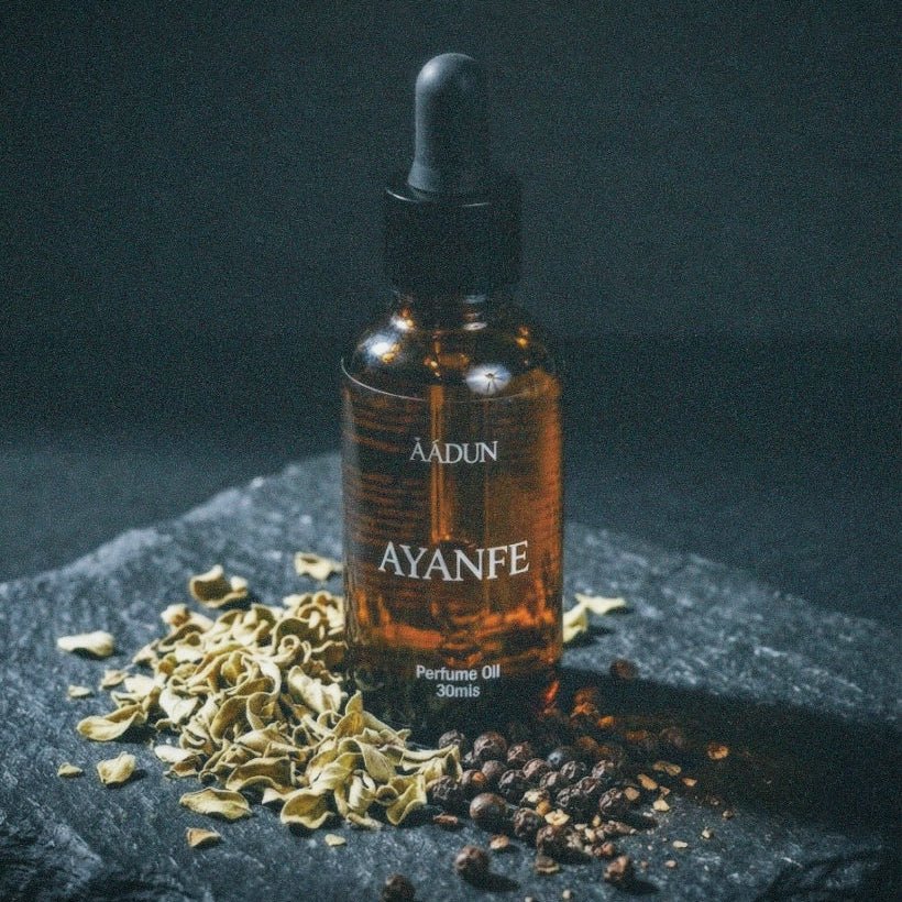 A 30ml amber glass dropper bottle of AADUN Ayanfe perfume oil, styled on a dark slate with dried botanicals and peppercorns against a dark background.