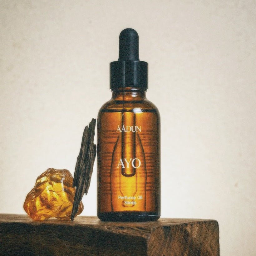 A small amber glass dropper bottle labeled AADUN AYO stands on a rustic wooden block against a warm beige textured background. A translucent amber crystal and dark dried botanicals are placed beside it as styled props.