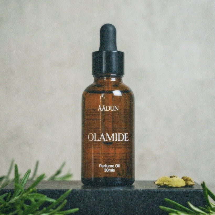 A 30ml amber glass dropper bottle of AADUN OLAMIDE Perfume Oil, styled on a dark stone block with rosemary, cardamom pods, and small red berries against a textured light gray background.