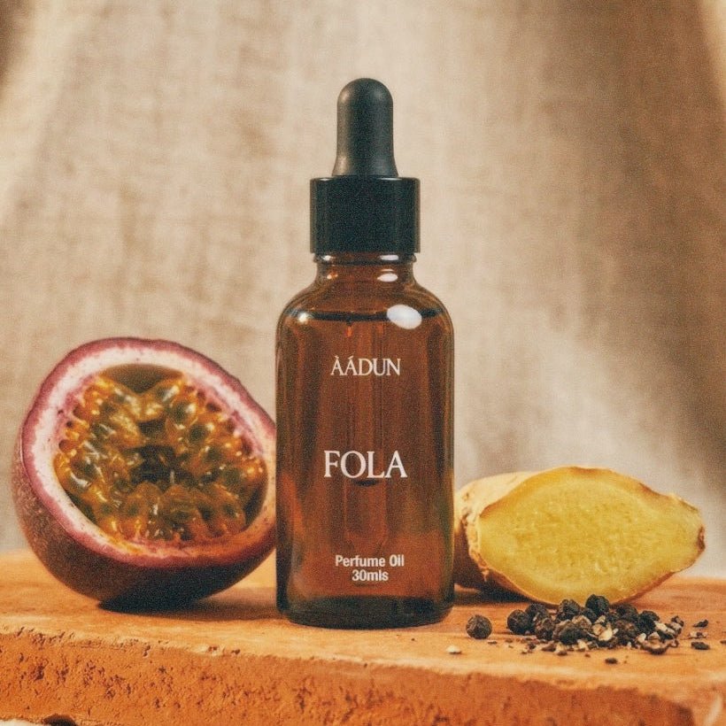 A bottle of AADUN FOLA perfume oil stands on a rustic wooden block, flanked by a halved passion fruit and a slice of ginger root against a draped beige fabric backdrop.