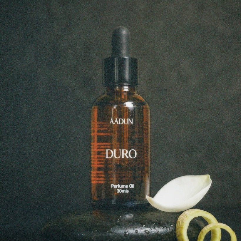 A bottle of AADUN DURO perfume oil stands on a dark stone surface against a dark gray background, styled with a white flower bud and yellow citrus peel slices.