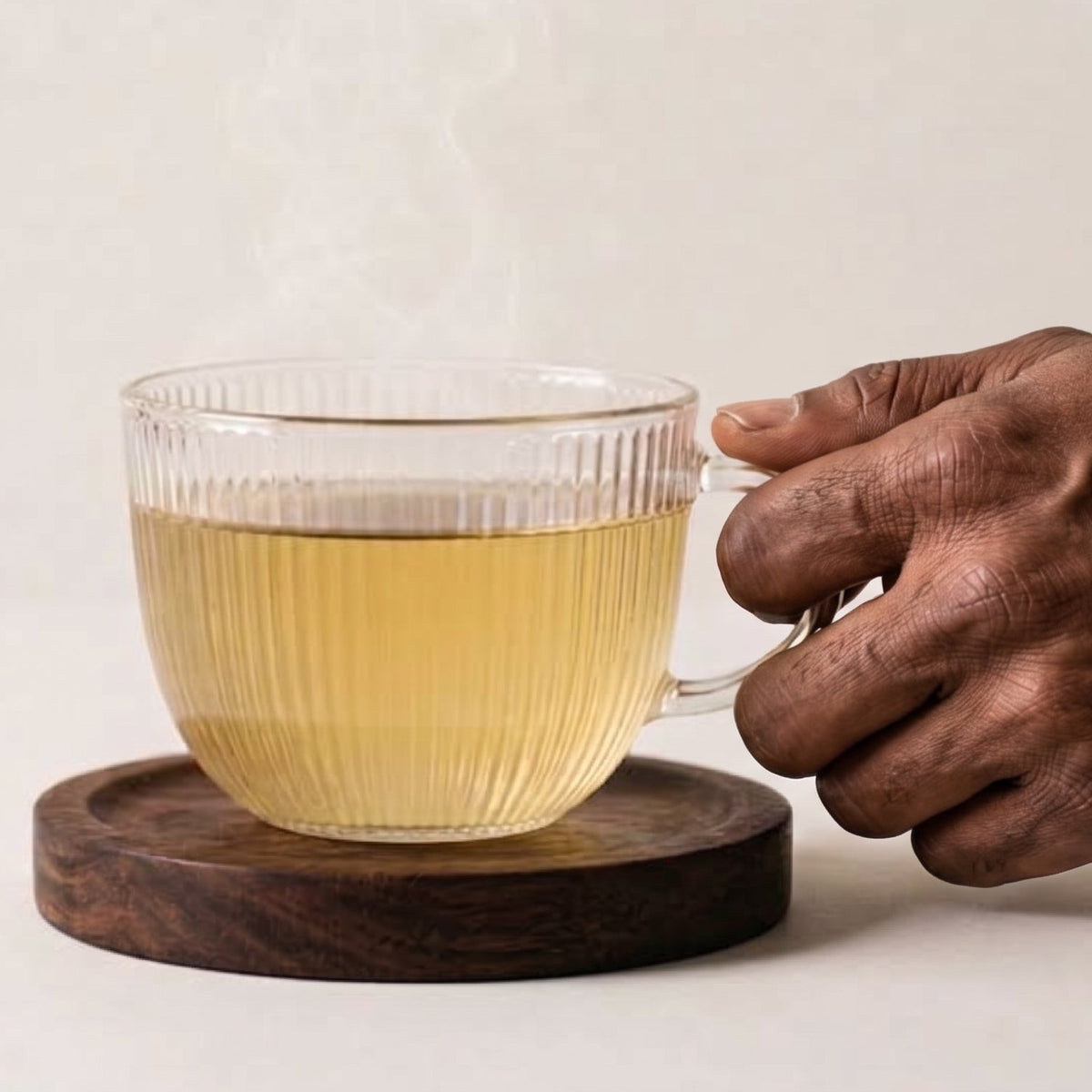 A clear ribbed glass mug filled with pale yellow tea, resting on a round dark wooden coaster, with a person's hand holding the handle and steam rising from the cup against a light neutral background.