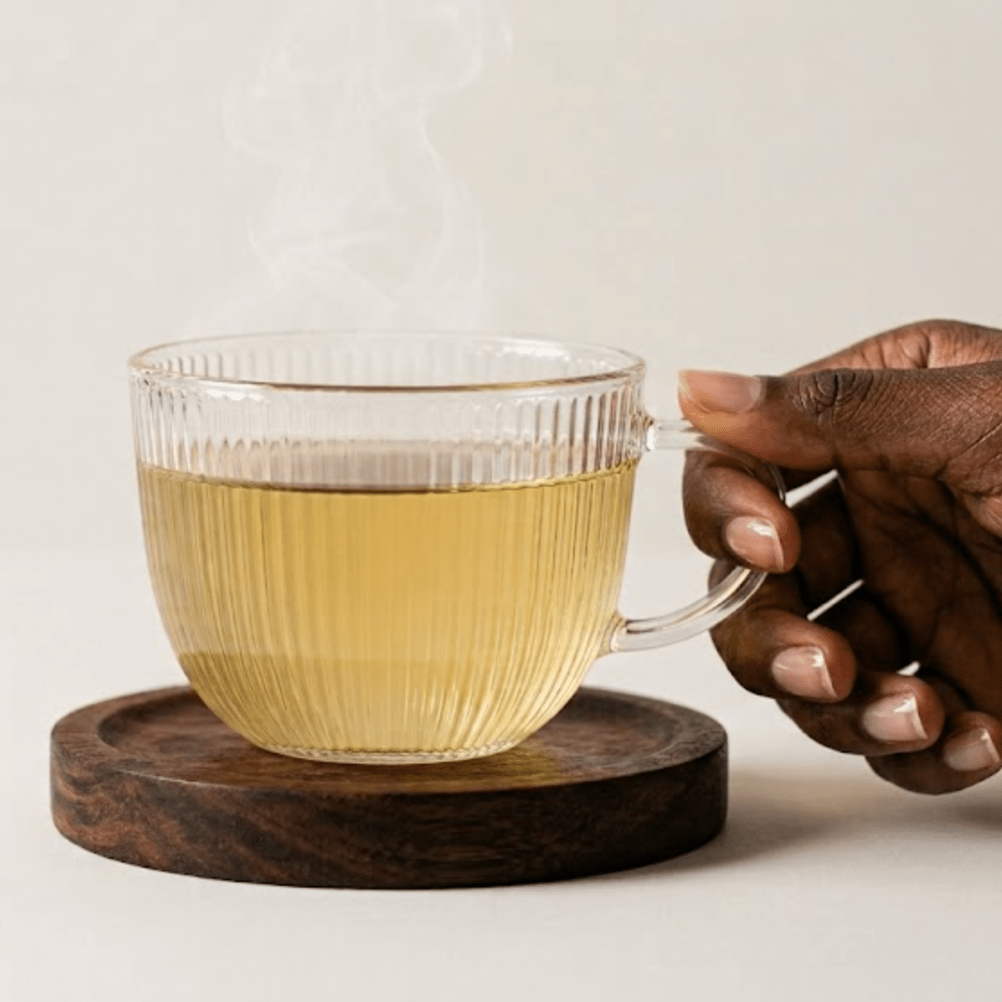 A clear ribbed glass mug filled with light amber tea sits on a round dark wooden coaster, with steam rising from the top. A person's hand holds the handle on the right side against a clean, warm off-white background.