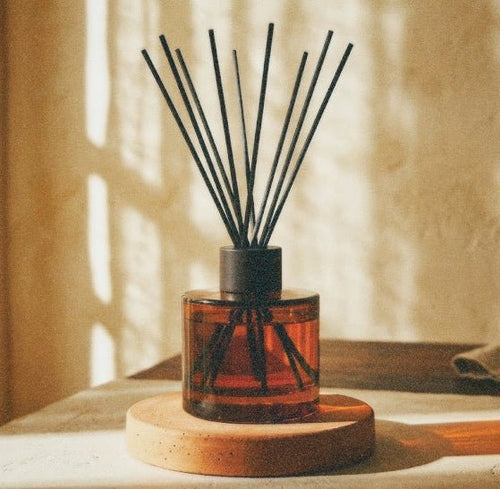 Amber glass reed diffuser bottle with black reeds on a round wooden base, placed on a table in warm sunlight with a textured wall and curtain in the background.