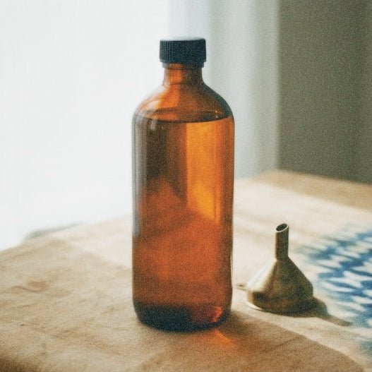 An amber glass bottle with a black cap stands on a wooden surface next to a small metal funnel, with a softly blurred indoor background.
