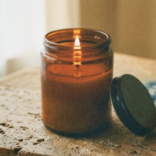 An amber glass jar candle with a lit flame sits on a textured stone surface, with its black lid resting beside it on a wooden tabletop.