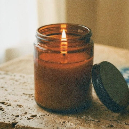 An amber glass jar candle with a lit flame sits on a textured stone surface, with its black lid resting beside it on a wooden tabletop.